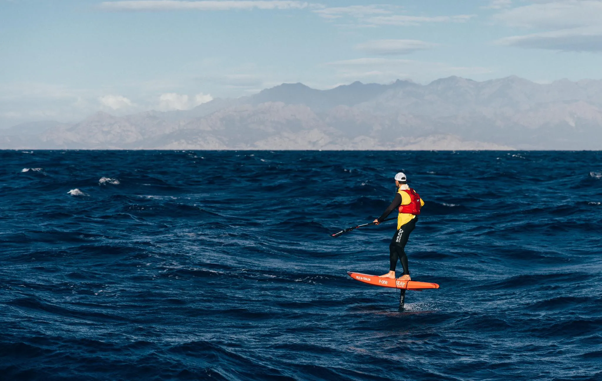 Erwan Jauffroy en SUP-foil, de dos, volant au-dessus de la houle avec sa pagaie en main lors d’un long downwind, une terre montagneuse visible en arrière-plan